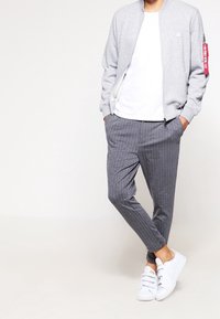 Grijze bomberjack met een ritssluiting aan de voorkant en een rood label, wit t-shirt, grijs gestreepte broek, en witte sneakers.