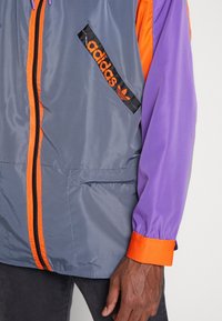 Grå Adidas-jacka med orange dragkedja och detaljer, lila ärmar, orange manschetter och en svart diagonal ficka med Adidas-logotyp på bröstet.