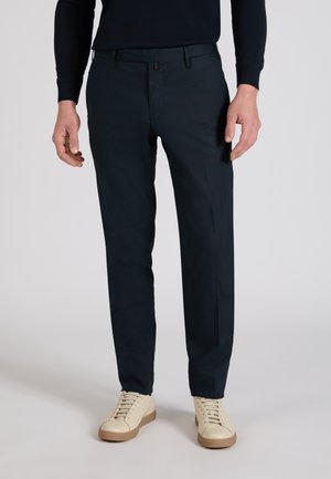 Mężczyzna w ciemnogranatowych, dopasowanych spodniach i beżowych tenisówkach na sznurowadła stoi na tle jednolitego jasnego tła.