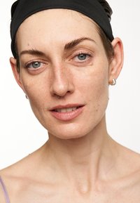 Close-up of a person with light skin, short hair covered by a black headband, light eyes, natural eyebrows, and silver hoop earrings.