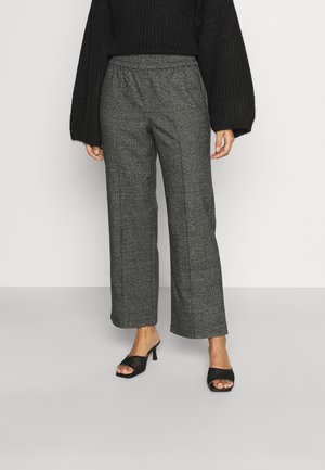 Femme portant un pantalon ample à carreaux gris, un pull en maille noir avec des manches ballon, et des sandales à talons ouvertes noires, debout contre un mur uni.