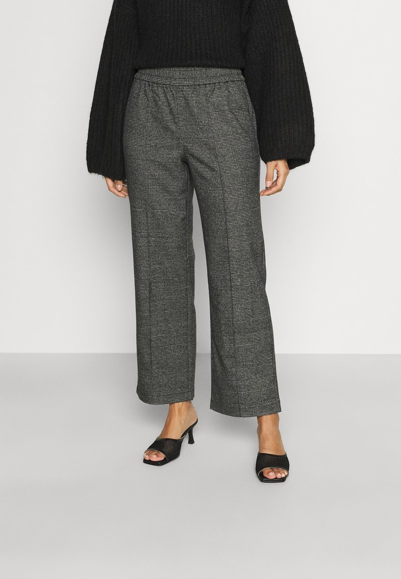 Femme portant un pantalon ample à carreaux gris, un pull en maille noir avec des manches ballon, et des sandales à talons ouvertes noires, debout contre un mur uni.