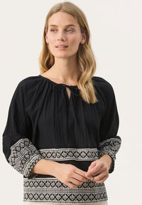 Zwarte blouse van een gestructureerde stof, met een geplooide halslijn, strikdetail en geborduurde witte patronen op de mouwen en de zoom.