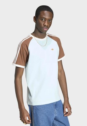 Ung mand iført en hvid og brun Adidas kortærmet skjorte og blå jeans, der står mod en ensartet lys grå baggrund.