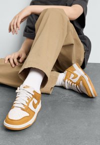 Nike tenisky v bielej farbe s akcentmi z pieskového semišu. Obsahujú biele šnúrky, perforovanú špičku a gumovú podrážku. Nosené s khaki nohavicami a bielymi ponožkami.