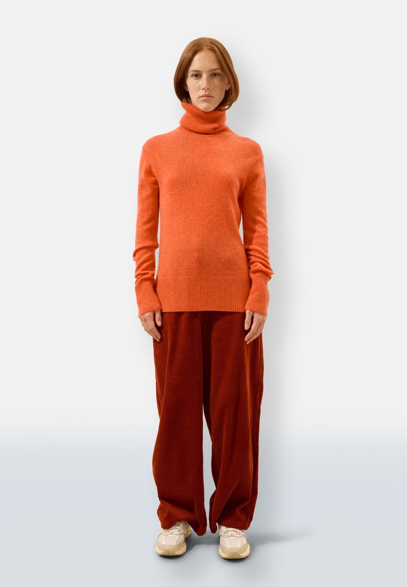 Oranges Rollkragenpullover mit gerippten Bündchen, kombiniert mit weiten Cordhosen in Rostfarbe. Weiße Sneakers runden das Outfit ab.