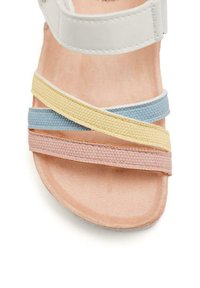 Sandalia con tiras de sujeción, con parte superior blanca y bandas cruzadas en azul, amarillo y rosa. Tiras de tela texturizada sobre una suela lisa de color marrón claro.