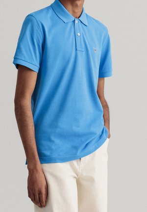 Homme portant un polo bleu vif à manches courtes et un pantalon beige clair, debout avec une main dans la poche devant un fond uni.