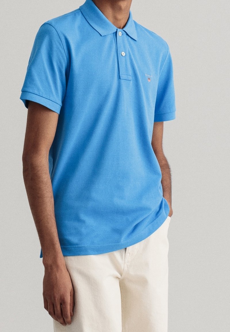 Homme portant un polo bleu vif à manches courtes et un pantalon beige clair, debout avec une main dans la poche devant un fond uni.