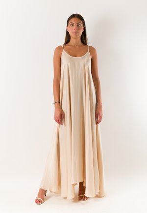 Donna in piedi che indossa un abito lungo beige fluido senza maniche e tacchi aperti, su uno sfondo chiaro e uniforme.