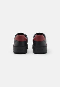 Černé kožené sneakers s burgundskými akcenty, kulatá špička, plochá gumová podrážka a embosované logo "BALLY" na patě. Hladký povrch a moderní design.