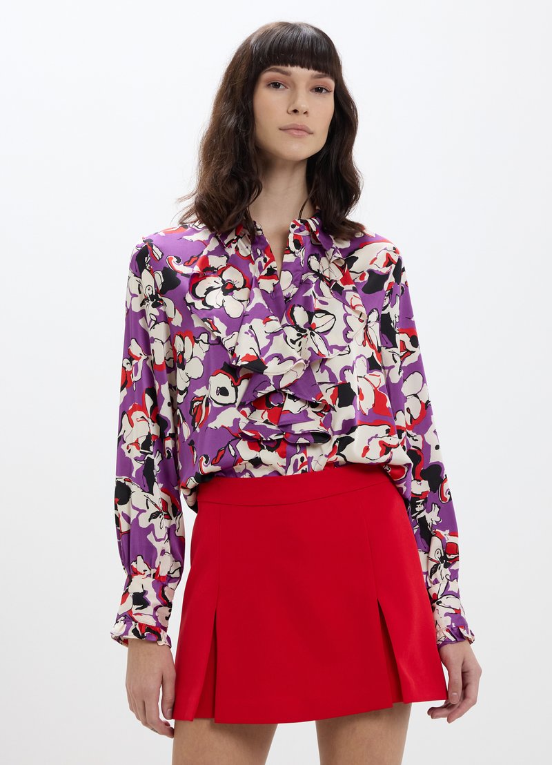 Blusa floreale viola con ruffles e maniche lunghe, abbinata a una gonna mini rossa con pieghe. Tessuto morbido e colori vivaci.