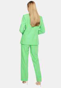 Jasnozielony garnitur składający się z dopasowanej marynarki i szerokich spodni, charakteryzujący się gładką fakturą i zorganizowanym fasonem. Widać eleganckie buty na obcasie.