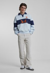 Lyseblå genser med marineblå striper, med oransje ørn-logo og ribbestrikkede mansjetter. Kombinert med kremfargede vide bukser og joggesko.