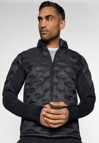 Μαύρο μπουφάν με επένδυση και φερμουάρ μπροστά, με επενδυμένο άνω μέρος και συμπαγείς μαύρες μανίκες. Περιλαμβάνει υποδοχές για τον αντίχειρα και λογότυπο στο στήθος.