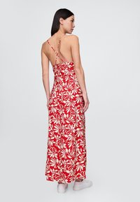 Rød maxikjole med blomsterprint, tætsiddende overdel, krydsede stropper og en flydende nederdel. Har store hvide blomsterprint og en rygfri design.