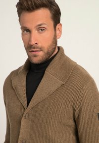 Brun strikket cardigan med et struktureret mønster, stor krave og knaplukning. Bæres over en sort turtleneck med et sort detaljer.