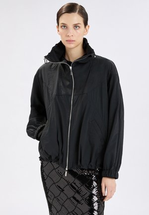 Donna con giacca nera con zip, collo alto e polsini elasticizzati, abbinata a una gonna nera con paillettes a motivo, in piedi davanti a uno sfondo bianco.