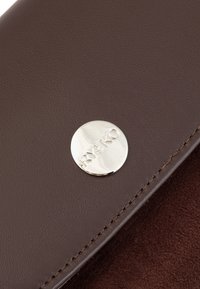 Brązowy skórzany produkt z okrągłym srebrnym logo i szczegółowym przeszyciem, prezentujący gładką teksturę oraz bogaty kontrast kolorystyczny.