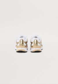 Buty sportowe z metalicznym złotym tyłem, białą siateczkową cholewką, czarną podeszwą i białymi detalami. Zaokrąglony kształt z minimalistycznymi akcentami.