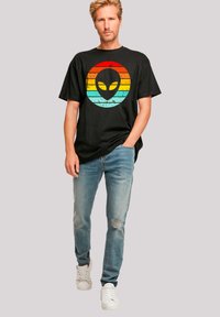 Schwarzes Baumwoll-T-Shirt mit einem verwitterten, mehrfarbigen Alien-Logo auf der Vorderseite, kombiniert mit hellblauen, verwitterten Jeans und weißen Turnschuhen.