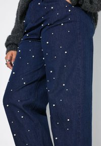 Dunkelblaue Jeans mit verstreuten Perlenakzenten. Der Schnitt ist lässig, mit klassischem Design und strukturierten Oberflächen.