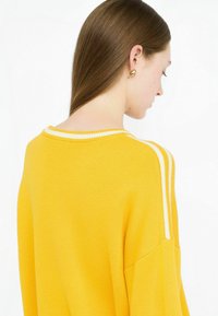 Pullover giallo a maglia con strisce bianche lungo il colletto e le maniche. Texture morbida, vestibilità rilassata e design con spalle abbassate.