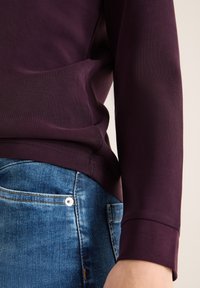 Lila langärmliges Oberteil mit einer weichen Textur; kombiniert mit blauen Denim-Jeans, die einen silbernen Knopf und kontrastierende Nähte aufweisen.