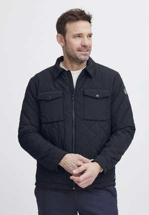 Schwarze gesteppte Jacke mit Reißverschluss, zwei Brusttaschen und spitzem Kragen. Strukturierter Stoff und sichtbare Nähdetails.