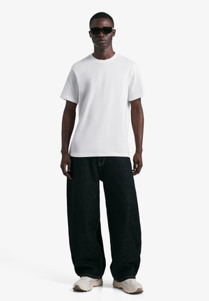 Bela bombažna majica s kratkimi rokavi, kombinirana s črnimi širokimi kavbojkami in nevtralnimi športnimi čevlji, nošena s temi sončnimi očali. Enostavna in moderna zasnova.