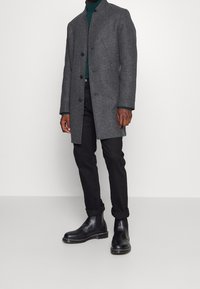 Cappotto in misto lana grigio con colletto alto, chiusura a bottoni, abbinato a pantaloni slim-fit neri e stivali Chelsea in pelle nera.