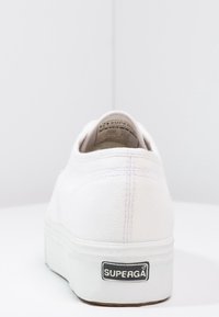 Baskets en toile blanche avec une surface texturée, un talon rembourré et une semelle en caoutchouc portant un logo Superga noir à l'arrière.