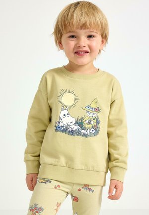Olivově zelená mikina s motivem Moomina, zobrazující slunce a dvě postavy mezi květinami. Doplněná vzorovanými krémovými legínami.