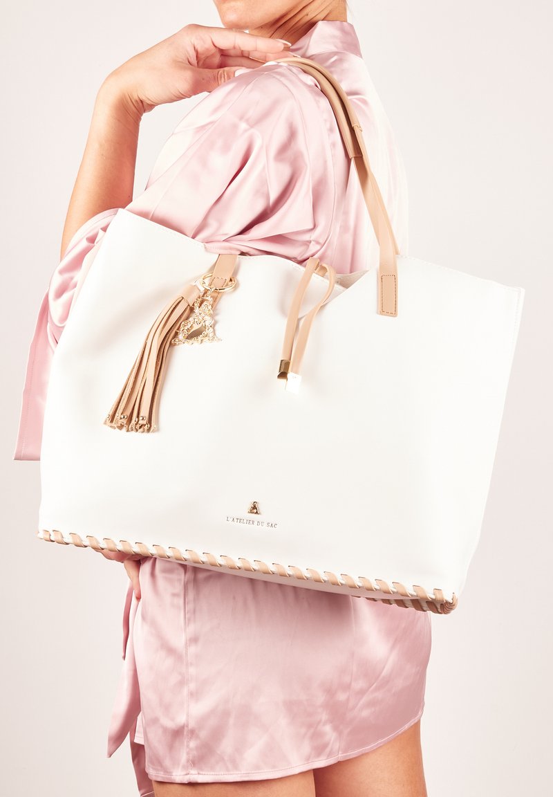 Biała torebka na ramię z beżowymi skórzanymi uchwytami, złotymi elementami i akcentem w postaci frędzla. Posiada wyszywane detale wzdłuż dolnej części.