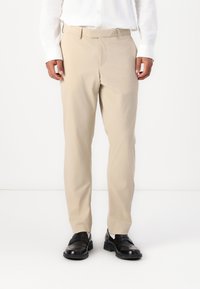 Pantaloni beige su misura con una superficie liscia, passanti per cintura e una piega frontale, abbinati a scarpe in pelle nere.