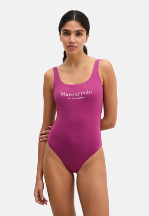 Frau mit zurückgebundenen dunklen Haaren, die einen ärmellosen magentafarbenen Badeanzug mit dem Schriftzug "Marc O'Polo" trägt und vor einem schlichten hellen Hintergrund steht.