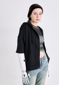 Crna zip-up dukserica kratkih rukava, uparena siva cropped majica i plave hlače s vertikalnim prugama. Uključuje protezu za ruku.