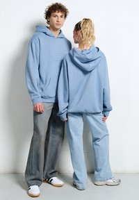 Młody mężczyzna zwrócony przodem i młoda kobieta tyłem, oboje ubrani w jasnoniebieskie bluzy z kapturem i luźne jeansy, stojący na tle białej ściany.