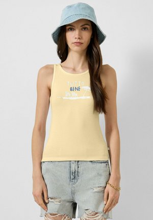 Femme portant un chapeau bob bleu clair, un débardeur jaune avec le texte « TUTTO BENE » et un motif plage, ainsi qu'un short en jean délavé clair.