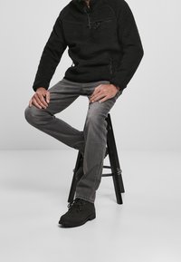 Schwarzer Fleece-Pullover, graue Jeans und schwarze Schnürstiefel. Der Pullover hat einen halben Reißverschluss-Kragen und ein strukturiertes Gewebe. Die Hocker haben eine runde Basis.