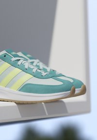 Paar türkisfarbene und hellgelbe Adidas-Sneaker mit weißen Sohlen auf einer weißen Oberfläche vor einem schattierten grauen und blauen Hintergrund.