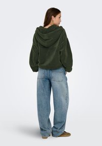 Sudadera de forro polar verde con capucha, corte holgado, gran bolsillo frontal, combinada con jeans azules sueltos y zapatillas tipo mocasín con estampado.