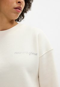 Krémová mikina s volným střihem, vyrobená z texturované látky, s nápisem "nová éra mě" obšitým krystaly na hrudi.