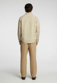 Camicia beige a maniche lunghe con orlo arrotondato e polsini abbottonati, abbinata a pantaloni larghi di un beige chiaro e scarpe nere.