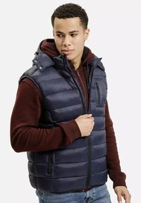 Młody mężczyzna noszący granatową ocieplaną kamizelkę na bordowej bluzie z kapturem, zapinający kamizelkę, patrzy w bok.