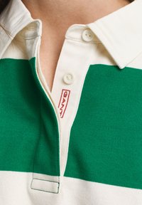 Grønn og hvit stripet polo skjorte med klassisk krage, plastknapper og et rødt "GANT" merke nær knappestolpen.
