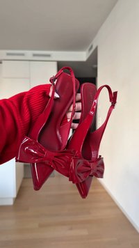 Red patent leather slingback heels feature a pointed toe and large decorative bow at the front, with a sleek ankle strap design.