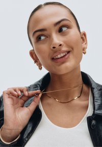 Kultainen kaulakoru, jossa on kaareva muotoilu ja tumma koriste. Malli käyttää myös kultaisia rengas- ja nappikorvakoruja sekä teksturoitua mustaa nahkatakkia.