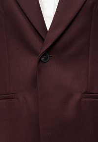 Μπορντό σακάκι από υφασμάτινη υφή, με γιακά με σχισμή, κλείσιμο με ένα κουμπί και δύο μπροστινές τσέπες. Φαίνεται η λεία φόδρα.