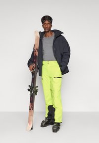 Άνδρας που φοράει μαύρη σκι jacket, γκρι πουκάμισο, φωσφοριζέ κίτρινα παντελόνια σκι, μπότες σκι και γυαλιά σκι, κρατάει ένα ζευγάρι σκι και στέκεται μπροστά από ένα απλό φόντο.